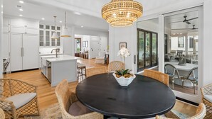 A modern kitchen with white cabinets, a central island, and a dining area with wicker chairs and a black table. - 8311 Jack Island Dr – 4 BR / Walk to Spring House / Elevator + Screened Porch (Johns Island)