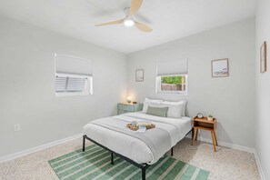 A bedroom with a bed, a bedside table, a nightstand, a ceiling fan, and two windows with blinds. - Beachside Boho Home. Fenced In Backyard & Short Walk to the Beach! 🌺🏖️ (Cape Canaveral)