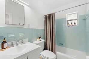 A bathroom with a white sink, two faucets, a toilet, a bathtub, a shower curtain, and a window with toiletries. - Beachside Boho Home. Fenced In Backyard & Short Walk to the Beach! 🌺🏖️ (Cape Canaveral)