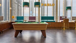 Un tavolo da biliardo in una stanza moderna con ampie finestre, lampade pendenti verdi e vista sulla città.