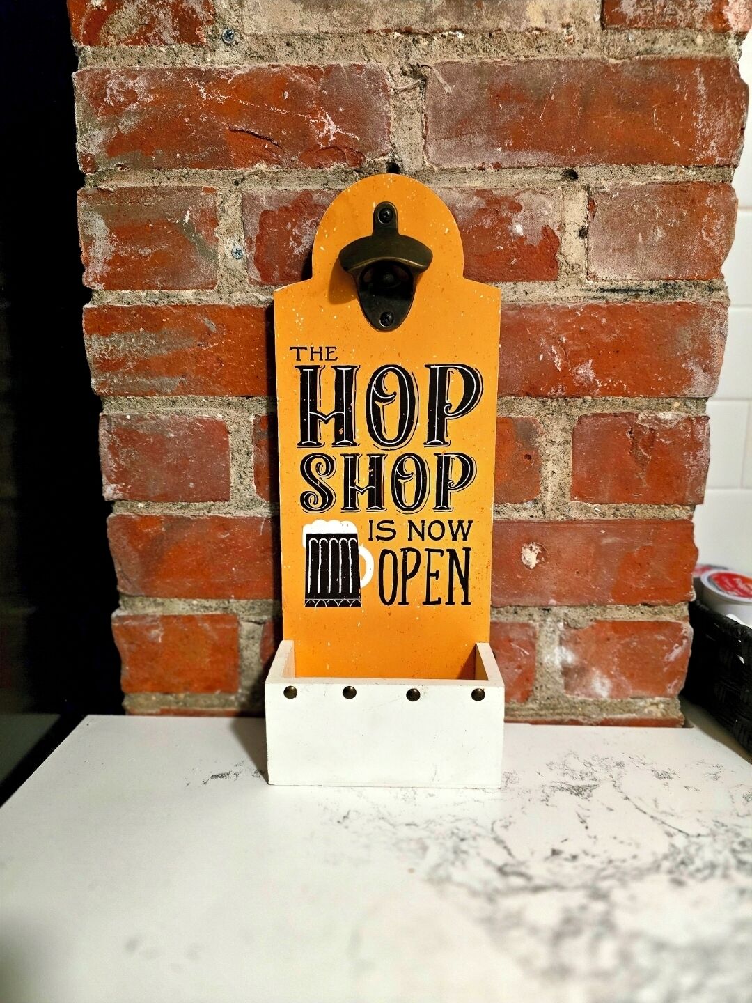 A sign with 'The Hop Shop is now open' against a brick wall backdrop.