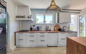 A kitchen with white cabinets, a gas stove, a coffee maker, and a hanging light fixture. - Charmante maison proche plage (Royan)