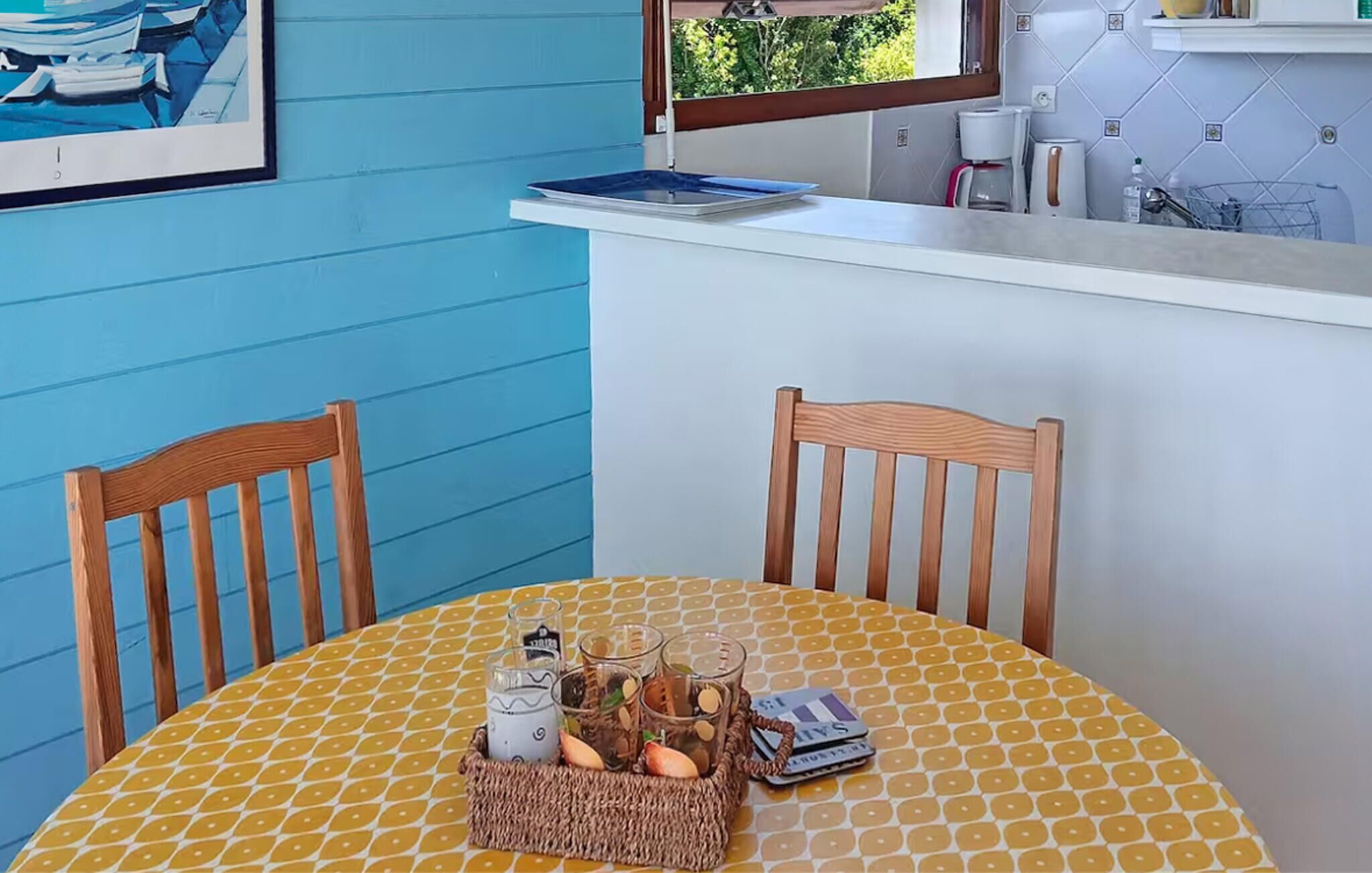 Un área de comedor con una mesa redonda, dos sillas de madera, una cesta tejida con vasos y una botella, y un ventanal con vista a vegetación.