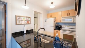 A modern kitchen with a black countertop, stainless steel sink, wooden cabinets, a refrigerator, and a microwave. - Lake Michigan Condo | Walk to Downtown | Views + Parking (Grand Haven)