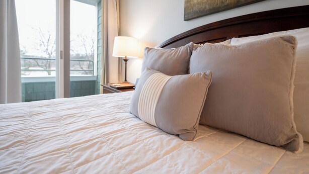 A hotel room with a large bed, a nightstand with a lamp, and a painting on the wall. - Lake Michigan Condo | Walk to Downtown | Views + Parking (Grand Haven)