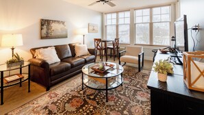 A living room with a leather sofa, a glass coffee table, a dining table with chairs, a ceiling fan, and a large window with blinds. - Lake Michigan Condo | Walk to Downtown | Views + Parking (Grand Haven)