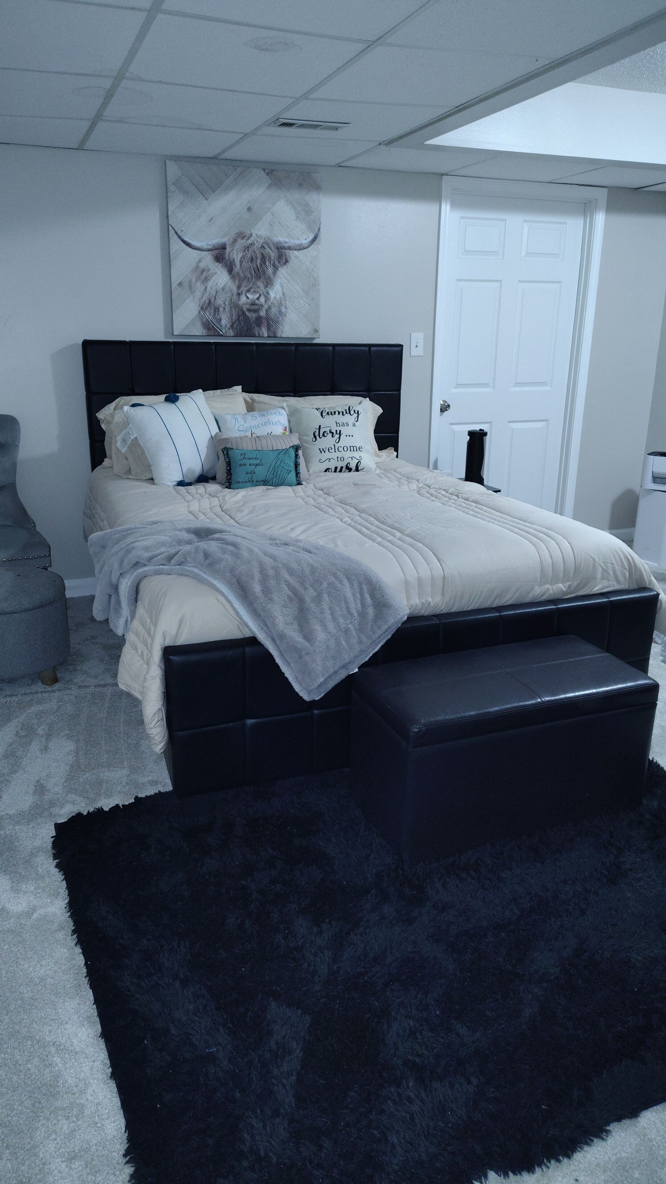 Une chambre à coucher avec un lit, des oreillers, un plaid, un pouf noir, un tapis noir, une porte blanche et un plafond muni d’une grille d’aération.