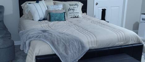Une chambre à coucher avec un lit, des oreillers, un plaid, un pouf noir, un tapis noir, une porte blanche et un plafond muni d’une grille d’aération.