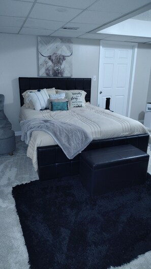 Ein Schlafzimmer mit Bett, Kissen, Tagesdecke, einem schwarzen Hocker, einem schwarzen Teppich, einer weißen Tür und einer Decke mit Lüftungsgitter.