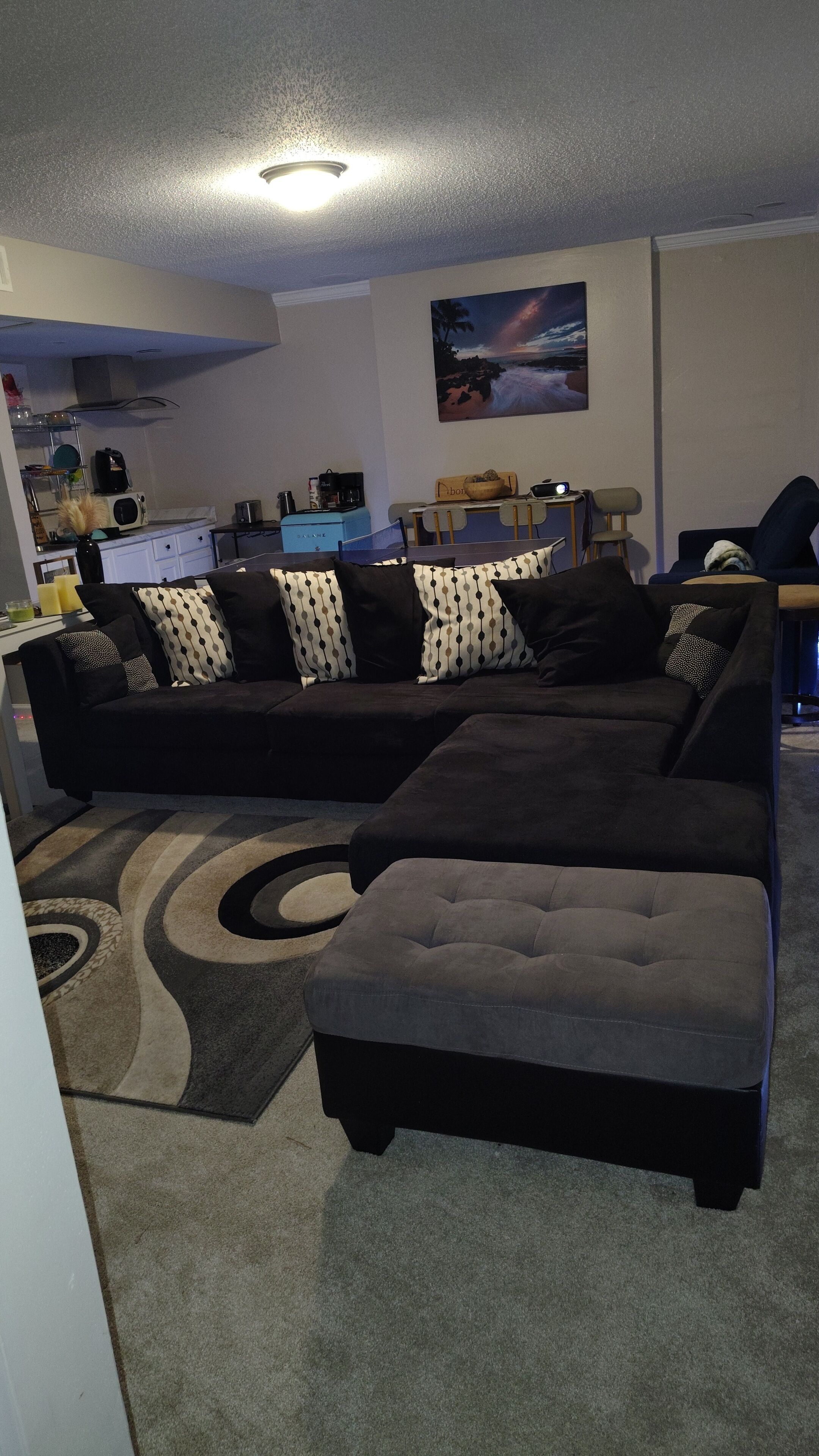 A living room with a sectional sofa, ottoman, and a painting on the wall.