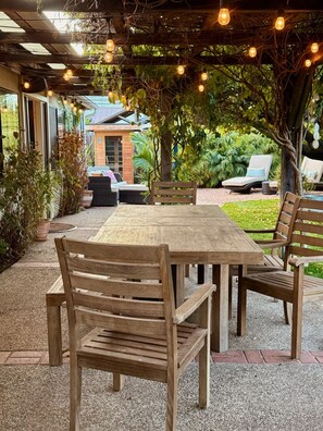 Une terrasse extérieure avec une table et des chaises en bois, entourée de verdure et agrémentée d’une pergola illuminée de guirlandes lumineuses.