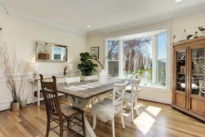 A dining room with a wooden table, white chairs, a large window with a view of trees, and a cabinet with glass doors. - This thoughtfully styled home overlooks the Bay and is perfect for your Annapolis escape. (Annapolis)