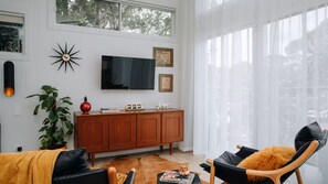 Una sala moderna con televisor de pantalla plana, aparador de madera, sillas de cuero y un ventanal con cortinas translúcidas.