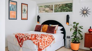Un dormitorio con cama, almohadas, una colcha, ventana, decoración en la pared, un reloj, una planta y un armario.