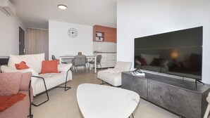 Ett modernt vardagsrum med en platt-TV, en matplats med stolar och ett kök i bakgrunden.