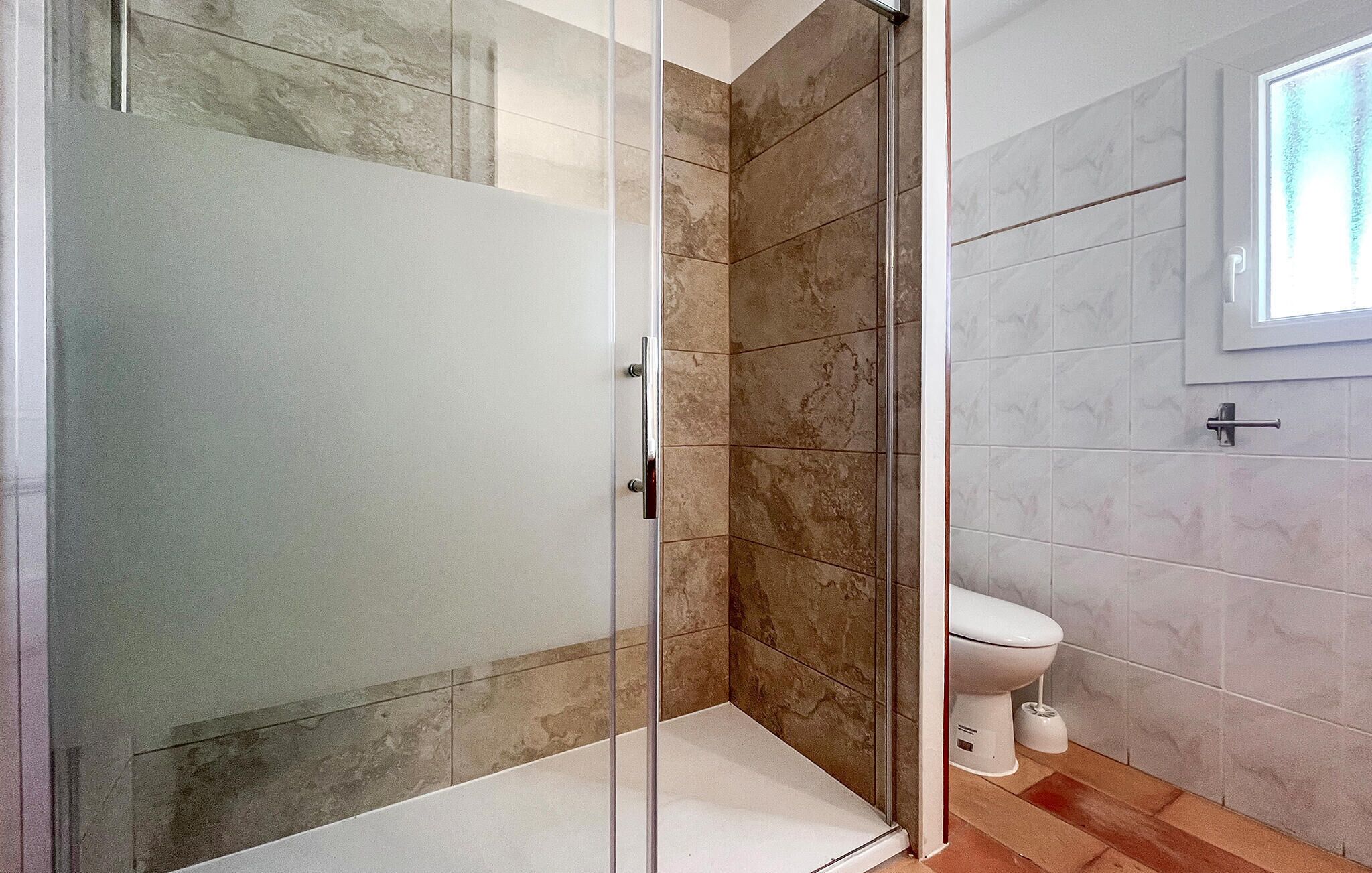 A bathroom with a glass shower enclosure, a toilet, and a window.