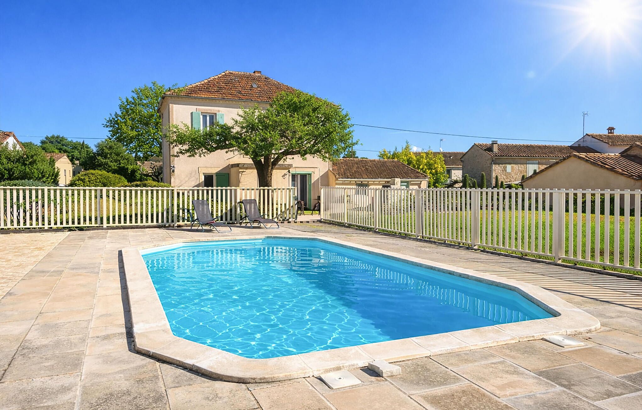 A rectangular swimming pool with a white fence and lounge chairs, surrounded by a house with a tiled roof and a tree.