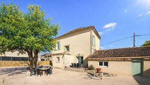 A two-story house with a tiled roof, a patio with a table and chairs, and a tree in the foreground. - Maison av. piscine proche Uzès (Saint Laurent la verne)