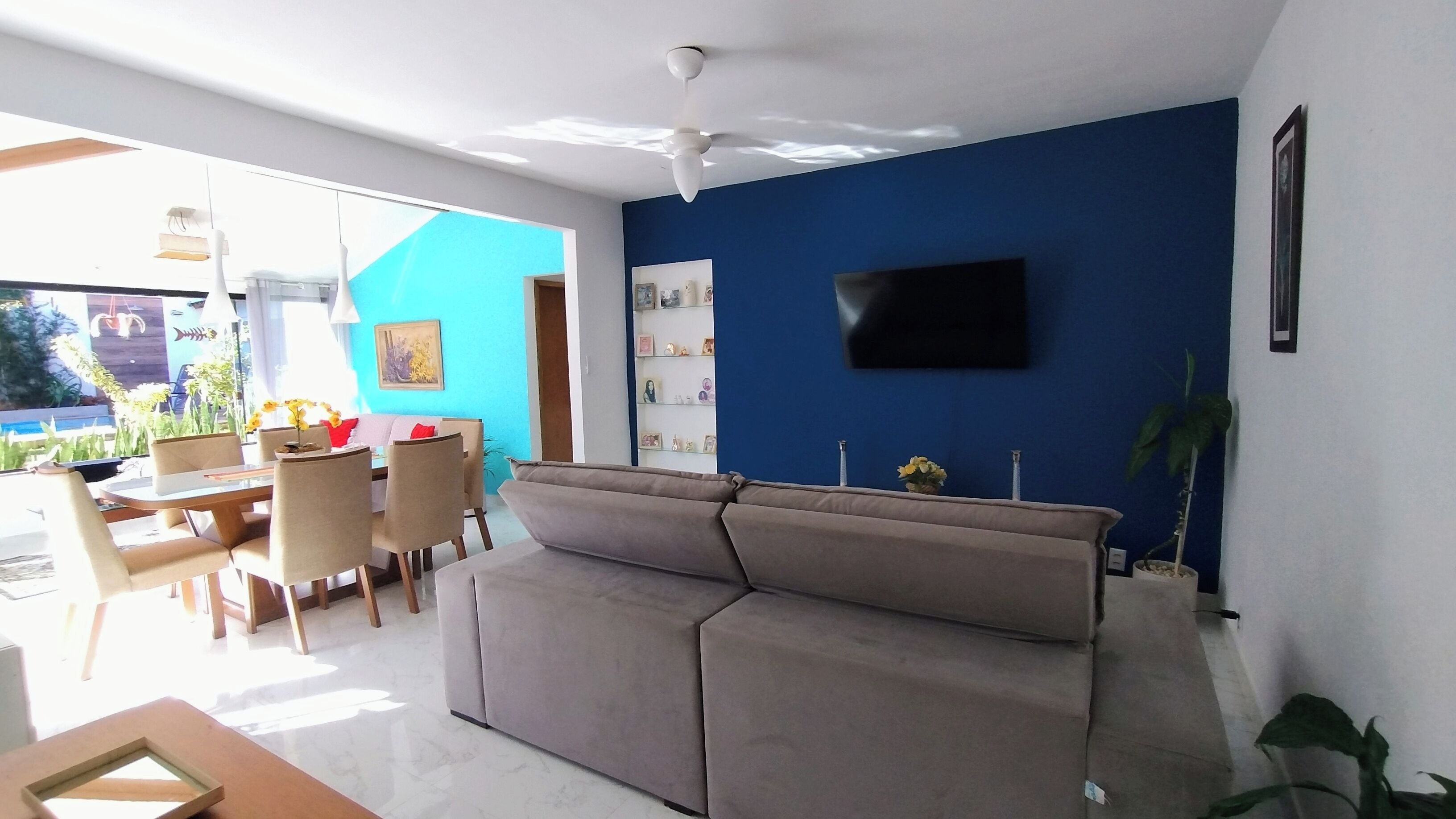 A living room with a sofa, dining table, chairs, a TV mounted on the wall, and a ceiling fan.