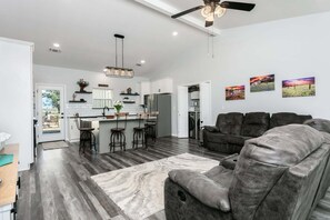 A spacious living area with a gray sofa, a kitchen with white cabinets, and a dining area with bar stools. - Ryder's Retreat | 2/2, Hot Tub, Fire-pit, Grill, EV Charger (Fredericksburg)