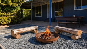 Una fogata con leña, grava y un edificio con ventanas y un porche cubierto al fondo.