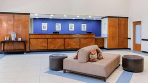 A reception area with a sofa, ottoman, and a desk with framed artwork. - La Quinta Inn & Suites Indianapolis Airport West (Plainfield)