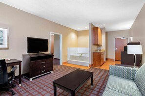 A hotel room with a flat-screen TV, a sofa, a desk with drawers, a small table, a bathtub, and a door leading to another room. - La Quinta Inn & Suites Indianapolis Airport West (Plainfield)