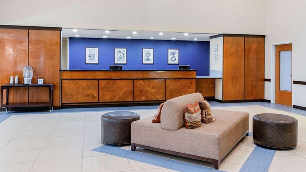 A reception area with a sofa, ottoman, and a desk with framed artwork. - La Quinta Inn & Suites Indianapolis Airport West (Plainfield)