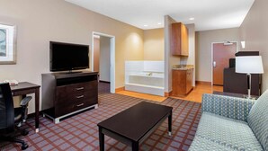 A hotel room with a flat-screen TV, a sofa, a desk with drawers, a small table, a bathtub, and a door leading to another room. - La Quinta Inn & Suites Indianapolis Airport West (Plainfield)