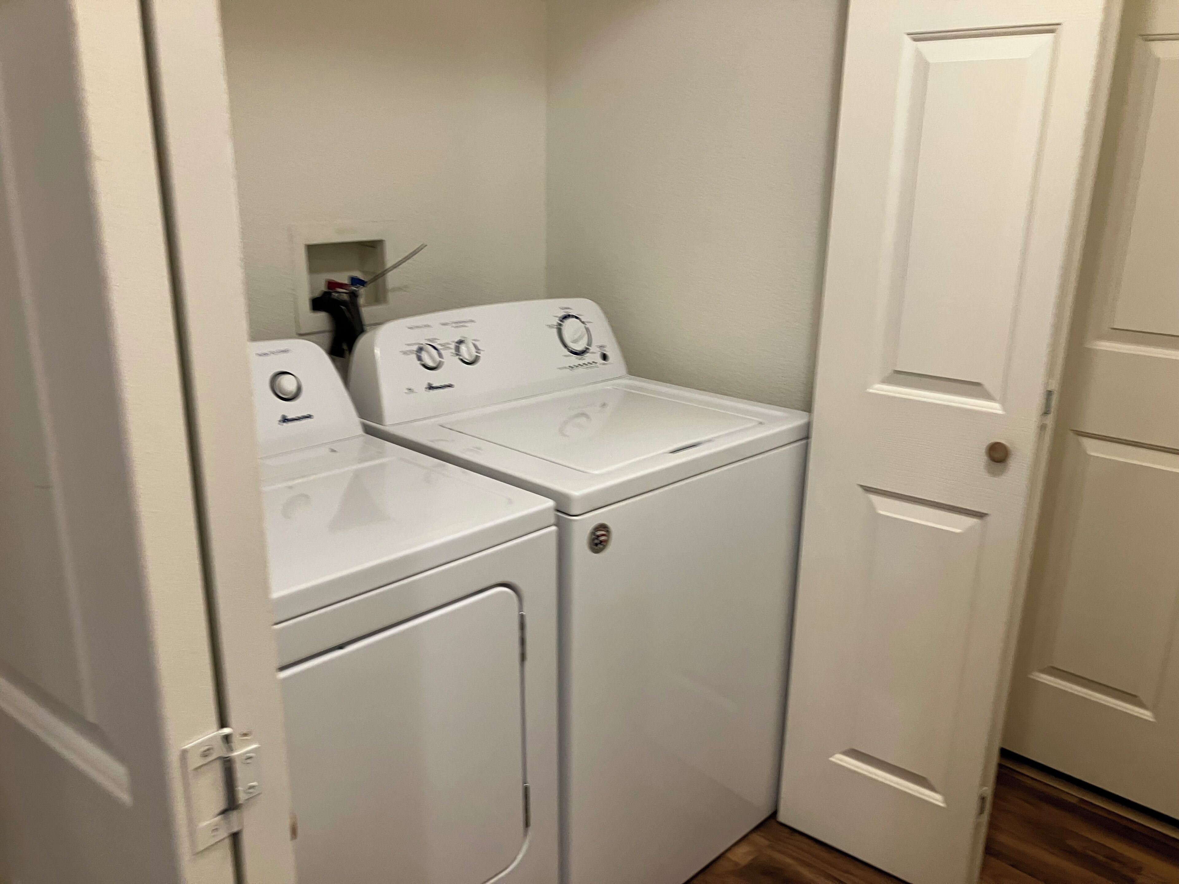 Une buanderie équipée de deux machines à laver et de deux sèche-linge à chargement frontal, tous deux de couleur blanche, d’une petite étagère contenant des produits de nettoyage et d’une porte située sur le côté droit.