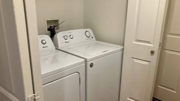 Une buanderie équipée de deux machines à laver et de deux sèche-linge à chargement frontal, tous deux de couleur blanche, d’une petite étagère contenant des produits de nettoyage et d’une porte située sur le côté droit.