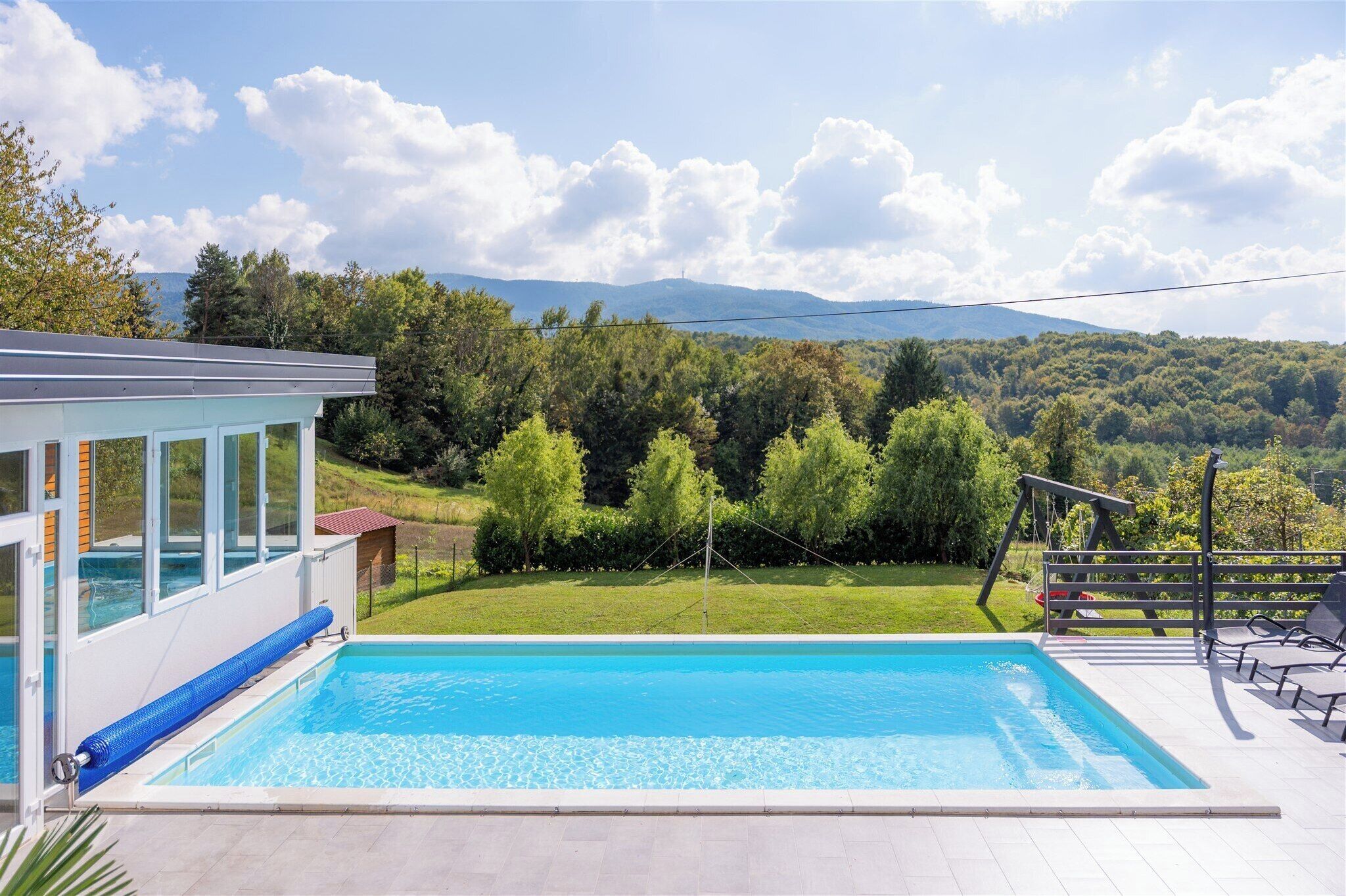 Une piscine avec vue sur une colline boisée, un espace couvert avec des sièges et un bâtiment doté de grandes fenêtres.