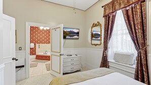 Een slaapkamer met een bed, een dressoir, een spiegel, een raam met gordijnen, een badkamer met een wastafel en een deur naar een andere kamer.