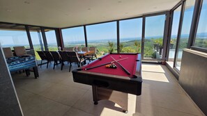 A pool table with a red felt surface, surrounded by chairs and a foosball table, with a view of a green landscape through large windows.