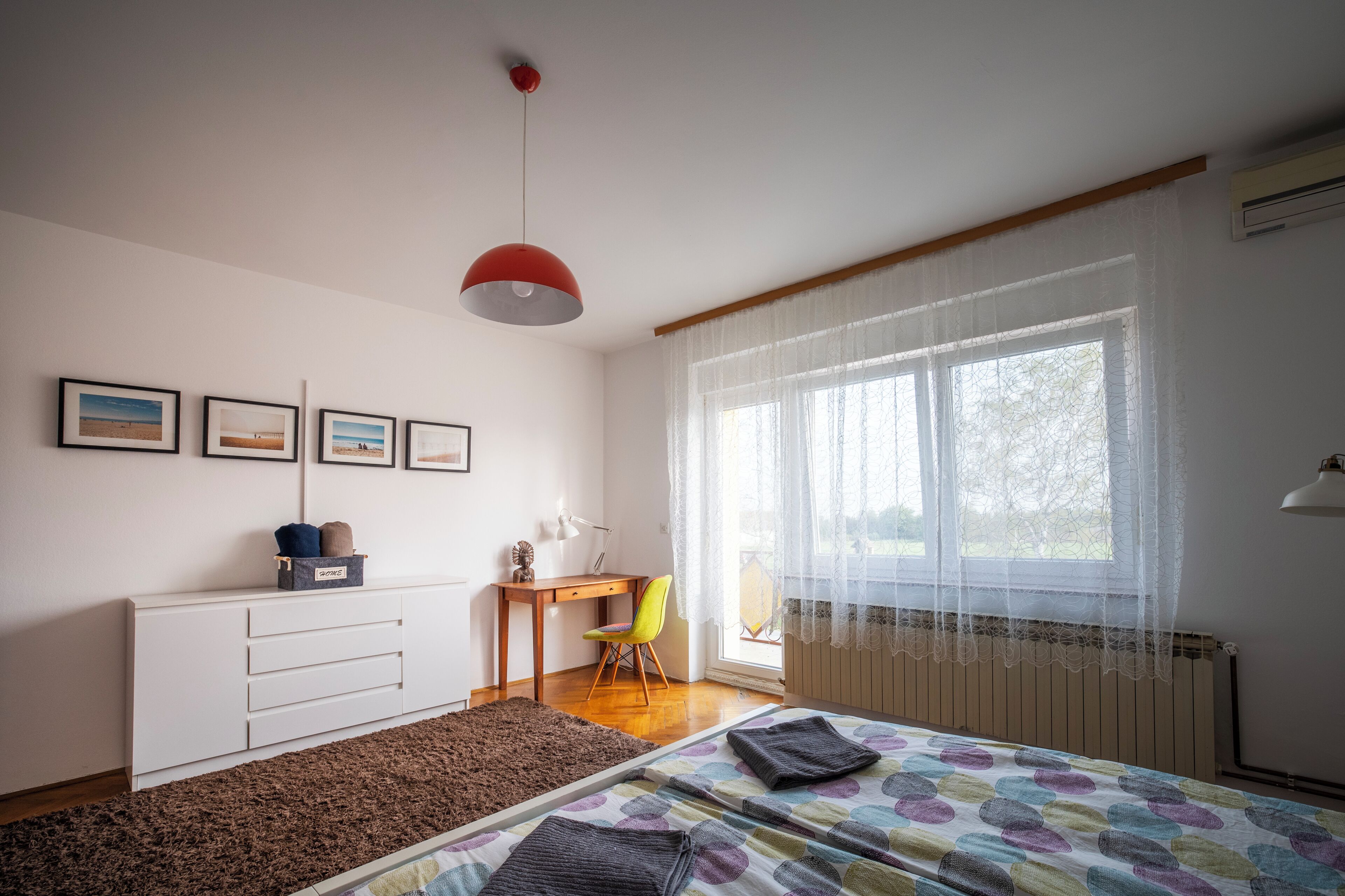 Un dormitorio con cama, escritorio, silla, radiador y ventana con cortinas traslúcidas.