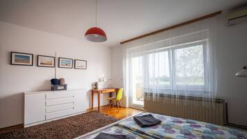 Un dormitorio con cama, escritorio, silla, radiador y ventana con cortinas traslúcidas.