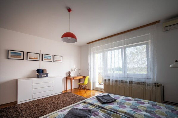 A bedroom with a bed, a desk, a chair, a radiator, and a window with sheer curtains.