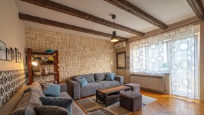 Un soggiorno con parete in pietra, travi in legno, un divano grigio, un tavolino in legno e tende con motivo decorativo.