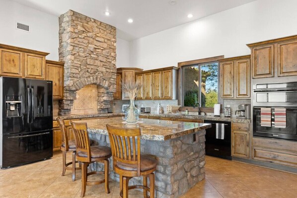 Una cocina con chimenea de piedra, armarios de madera, un refrigerador negro y una isla grande con sillas.