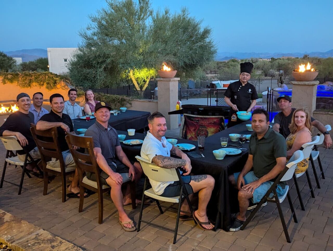A group of people seated at an outdoor dining table with a chef standing behind.