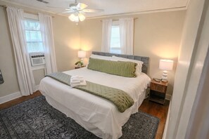 Una cama bien hecha con un almohadón verde, una lámpara de noche y un ventilador de techo en una habitación con un tapete estampado y una ventana con cortinas.