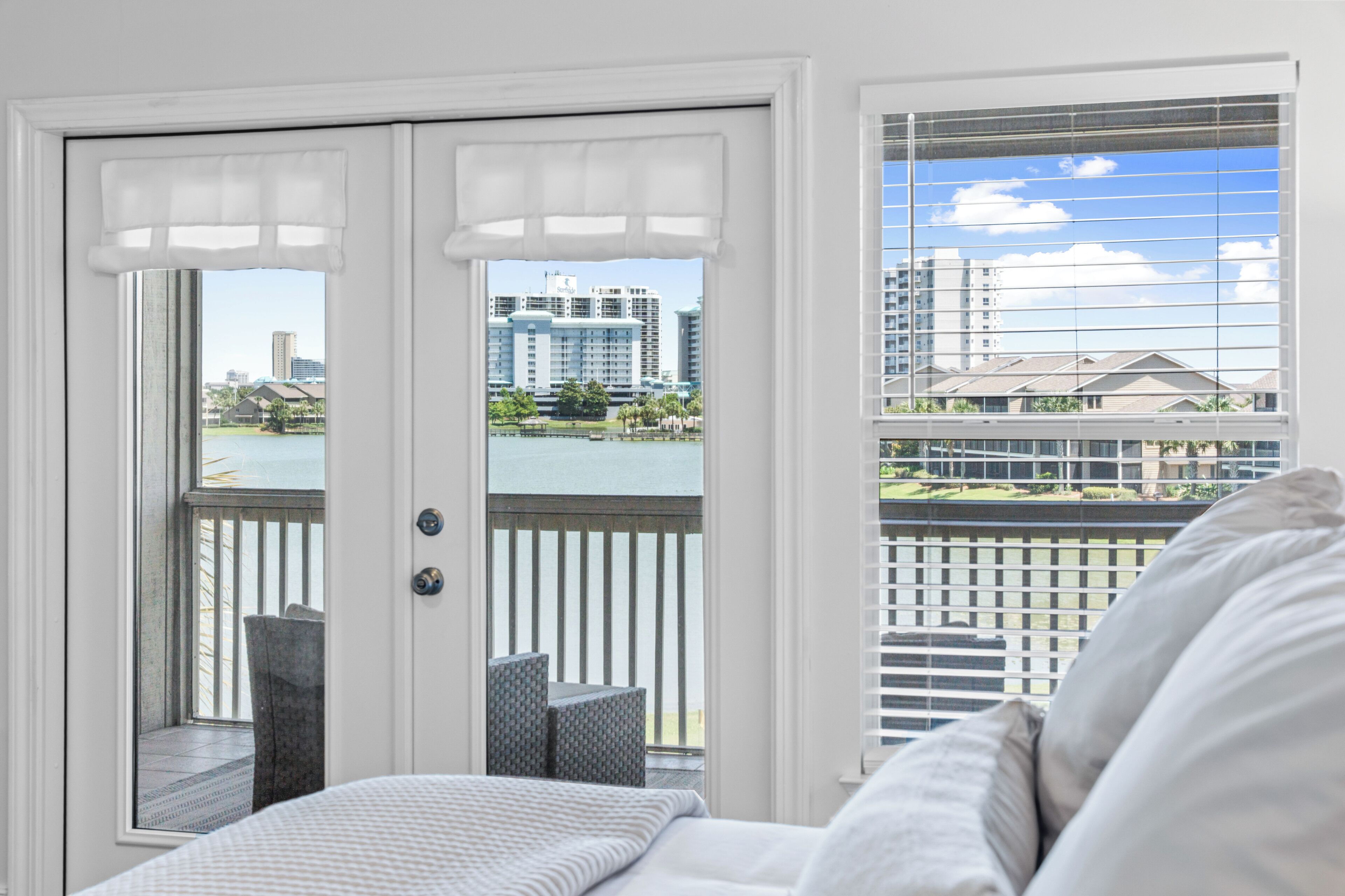 A room with a bed, a view of a balcony with a railing, and a door with a view of buildings and a body of water.