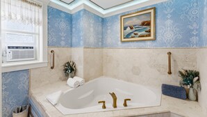 Une salle de bain avec une grande baignoire, une fenêtre ornée d’un rideau en dentelle et un tableau représentant un paysage côtier.