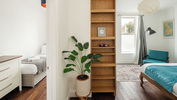 A bedroom with a bed, a wooden shelf unit, a potted plant, a map on the wall, and a window with curtains. - Bright 2BR Home with Private Patio (Greater London)