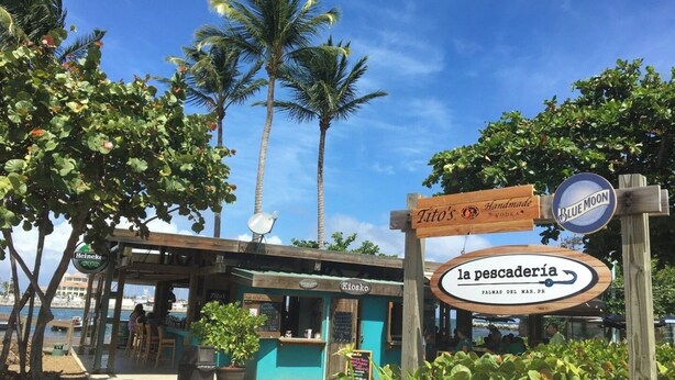 A beachfront restaurant with outdoor seating, a sign for 'la pescadería,' and palm trees in the background.