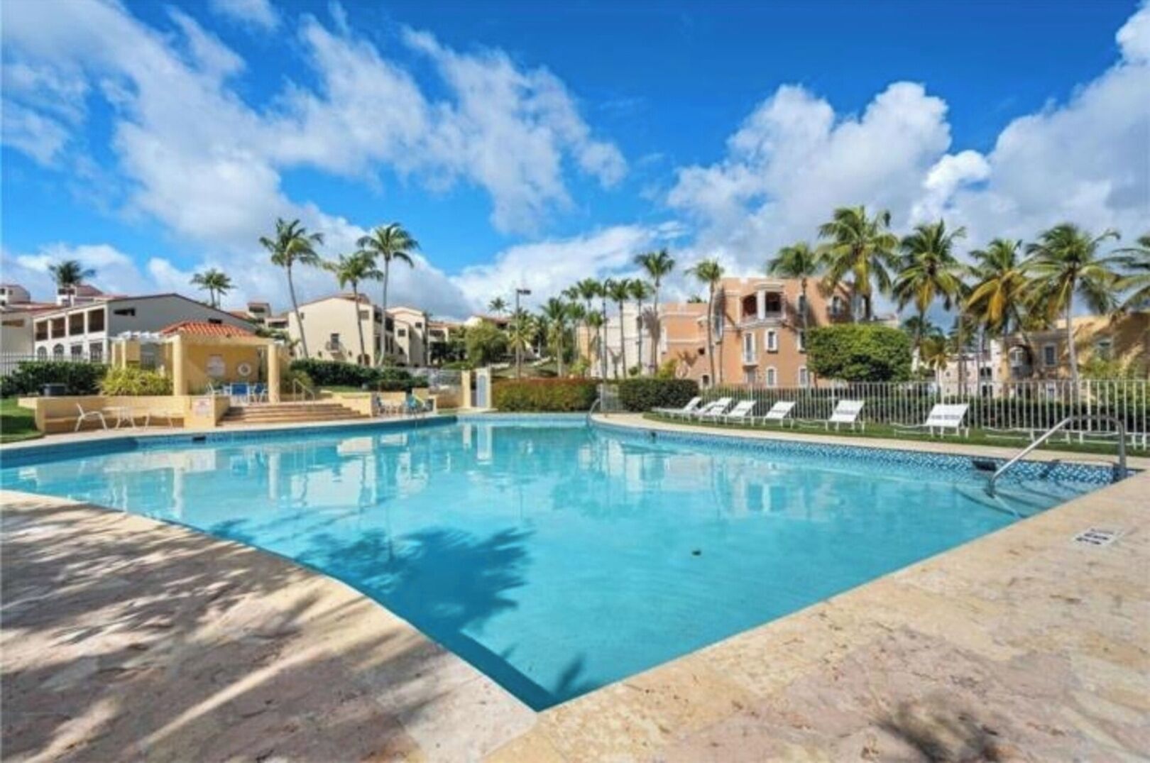 A swimming pool with lounge chairs, palm trees, and buildings in the background.
