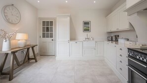 A kitchen with white cabinetry, a clock on the wall, a table with a lamp, and a stove. - The Lookout, Southwold (Southwold)