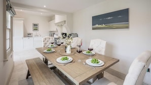 A dining area with a wooden table set for four, white upholstered chairs, and a painting on the wall. - The Lookout, Southwold (Southwold)