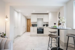 A modern kitchen with white cabinets, a dining area with stools, and a small table with a vase and wine. - The Vault, Southwold (Southwold)