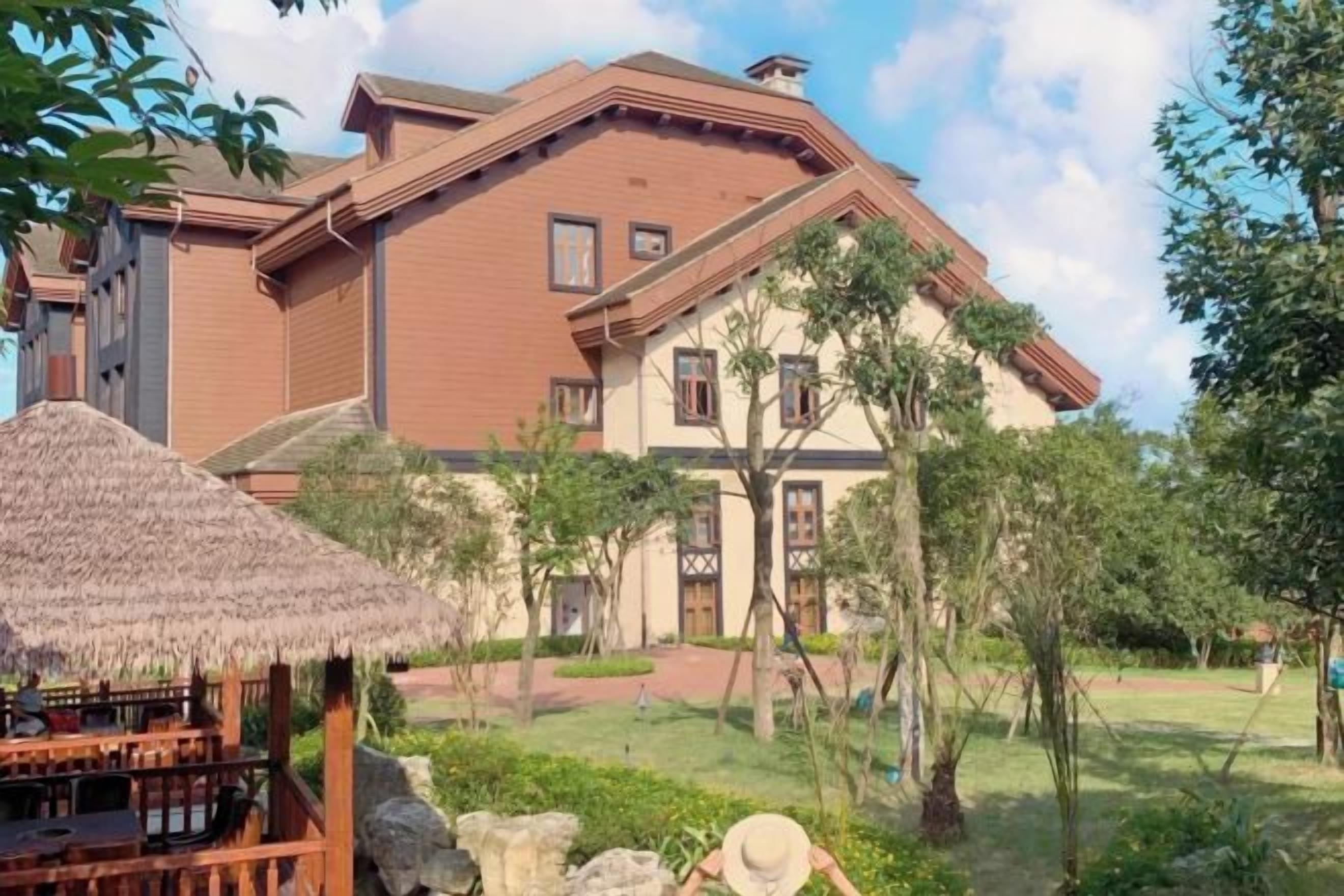 A two-story building with a thatched-roof structure in the foreground, surrounded by greenery.
